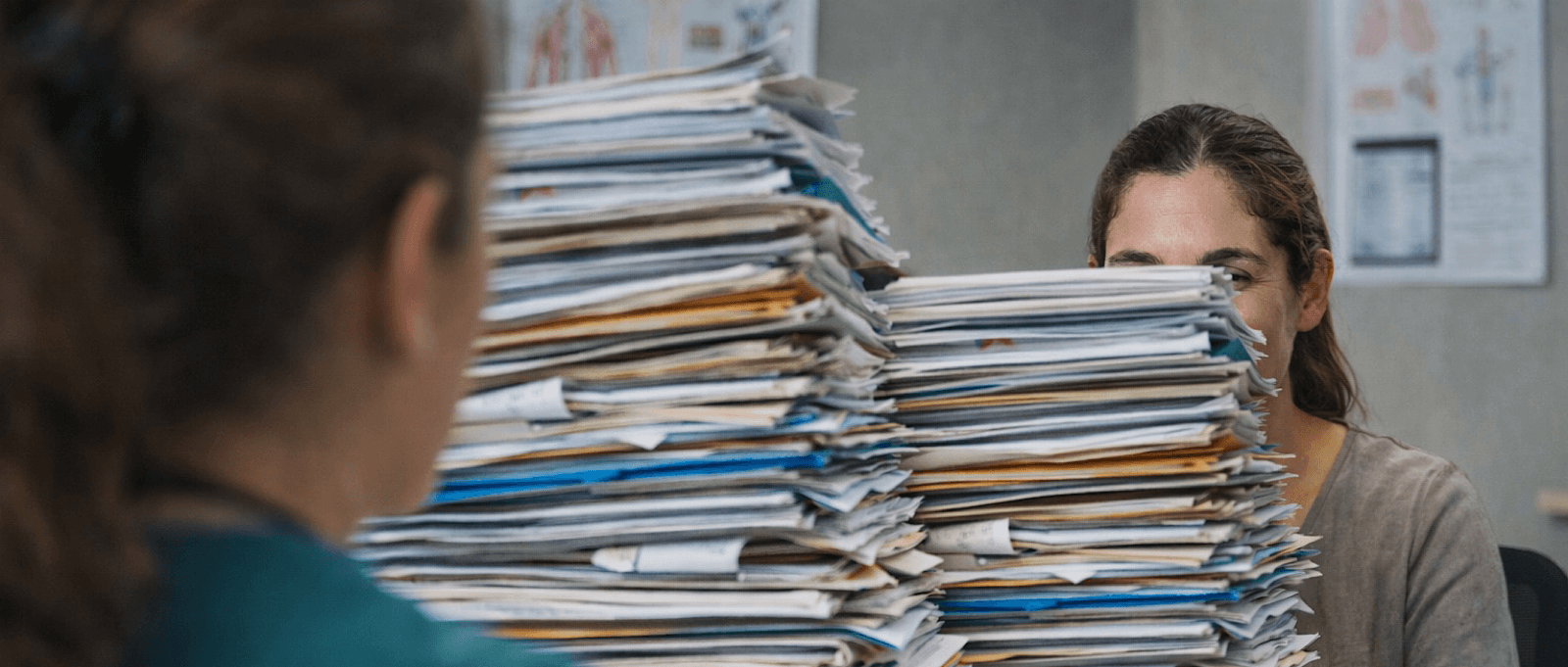 Patientin mit großem Stapel medizinischer Befunde sitzt gegenüber einer Internistin in der Ordination – Symbolbild für körperliche Beschwerden ohne erklärenden Befund.